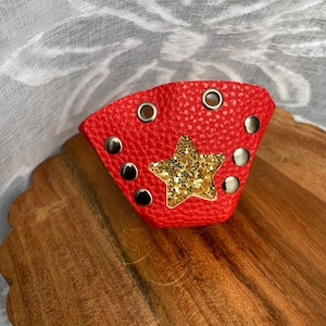 May include: A red leather wrist cuff with a gold glitter star in the center. The cuff has silver rivets and eyelets. The cuff is approximately 3 inches wide.