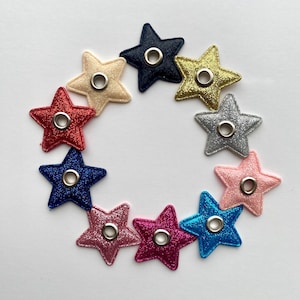 Puede incluir: Un conjunto de diez parches de tela con forma de estrella brillante con ojales en varios colores, incluidos rojo, rosa, azul, dorado y plateado.