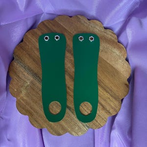 Puede incluir: Dos correas de zapatos de cuero verde con ojales metálicos. Las correas están sobre una superficie de madera.