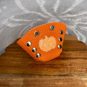 Op de afbeelding: Een oranje leren manchetarmband met zilveren klinknagels en oogjes. Een glinsterende pompoen is gecentreerd op de armband. De armband wordt gepresenteerd op een houten oppervlak.