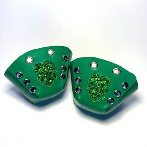 May include: A pair of bright green accessories, possibly shoe covers, with a glittery green leaf design. Each piece has multiple silver grommets and a white grommet. The items are set against a white background.