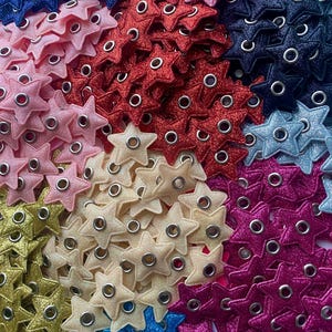 Puede incluir: Una colecci&oacute;n de adornos brillantes en forma de estrella con ojales met&aacute;licos en varios colores, incluyendo rosa, rojo, dorado, crema, azul y morado.