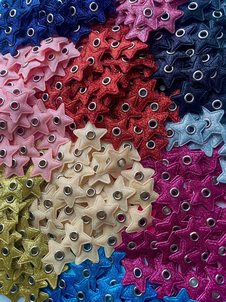 Puede incluir: Una colecci&oacute;n de adornos brillantes en forma de estrella con ojales met&aacute;licos en varios colores, incluyendo rosa, rojo, dorado, crema, azul y morado.