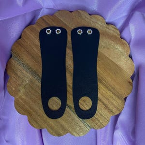 May include: Two black leather wrist straps with silver metal eyelets. The straps are on a wooden surface.