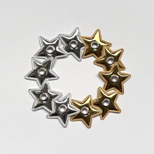 Puede incluir: Un conjunto de once ojales metálicos con forma de estrella dispuestos en un patrón circular. Cinco de los ojales tienen un acabado plateado brillante, mientras que los seis restantes presentan un acabado dorado pulido. Cada estrella tiene un orificio.
