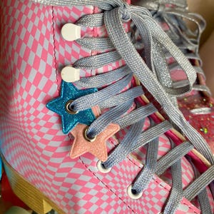 Puede incluir: Patines de ruedas a cuadros rosa y blancos con abalorios en forma de estrella azul y rosa en los cordones. Los cordones est&aacute;n atados en forma de cruz.
