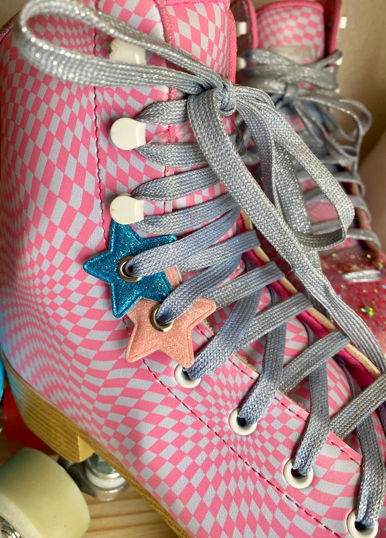 Puede incluir: Patines de ruedas a cuadros rosa y blancos con abalorios en forma de estrella azul y rosa en los cordones. Los cordones est&aacute;n atados en forma de cruz.