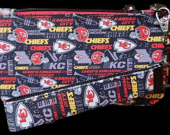 May include: A black zippered pouch with a black and white strap. The pouch and strap are covered in a repeating pattern of red, yellow, and white Kansas City Chiefs logos, text, and musical notes.
