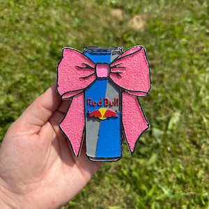 Könnte beinhalten: Ein Accessoire in Form einer Red Bull-Dose mit einer rosa Glitzer-Schleife. Die Dose zeigt das Red Bull-Logo und ist blau, grau und rot gefärbt. Die Schleife ist leuchtend rosa und hat eine glitzernde Textur.