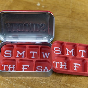 Puede incluir: Un organizador de pastillas rojo con una tapa plateada. La tapa tiene la palabra "VEIOD2" grabada en relieve. El organizador de pastillas tiene siete compartimentos etiquetados "SMTWT", "TH", "F", "SA", "TH", "F", y "SA".