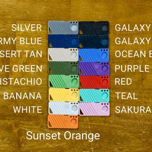 May include: A set of 12 colorful plastic keychains with different colors and patterns. The colors include silver, army blue, desert tan, olive green, pistachio, banana, white, galaxy black, galaxy blue, ocean blue, purple, red, teal, and sakura pink. The keychains are arranged in two rows of six, with the text "Sunset Orange" below the bottom row.