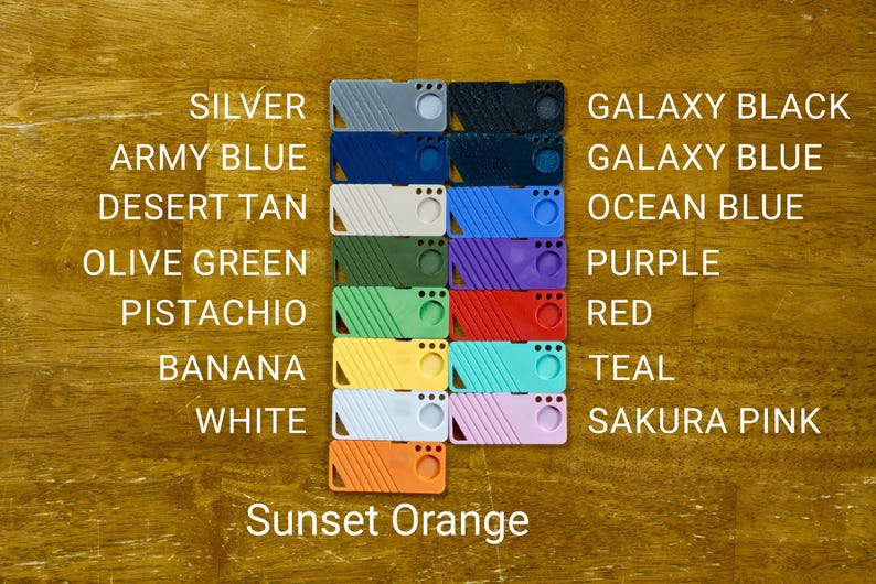 May include: A set of 12 colorful plastic keychains with different colors and patterns. The colors include silver, army blue, desert tan, olive green, pistachio, banana, white, galaxy black, galaxy blue, ocean blue, purple, red, teal, and sakura pink. The keychains are arranged in two rows of six, with the text "Sunset Orange" below the bottom row.
