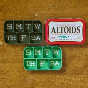 May include: Two green pill organizers with the days of the week labeled. The top organizer has a dark green background and white lettering. The bottom organizer has a light green background and white lettering. A red tin of Altoids mints is in the center of the image.