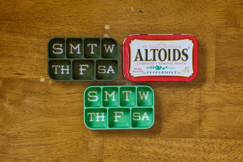 May include: Two green pill organizers with the days of the week labeled. The top organizer has a dark green background and white lettering. The bottom organizer has a light green background and white lettering. A red tin of Altoids mints is in the center of the image.