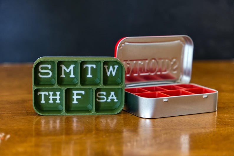May include: A green pill organizer with the days of the week labeled: S M T W TH F SA. A silver pill box with red compartments is also shown.