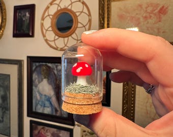 May include: A miniature red and white mushroom figurine with a spotted cap, resting on a patch of green moss inside a small clear glass dome with a cork base. The glass dome is cylindrical with a rounded top, enclosing the mushroom and moss.