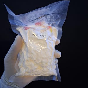 May include: A clear plastic bag filled with white, textured candy, labeled "Peach Rings." The bag is held by a gloved hand against a black background. Some peach-colored candy pieces are visible.