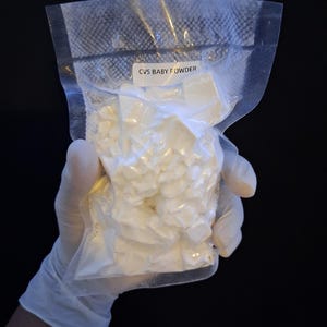 May include: A clear plastic bag filled with white powder, labeled "CVS BABY POWDER." The bag is held by a gloved hand against a black background. The powder appears to be in small, irregular clumps.