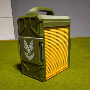 May include: An olive green box with a yellow slatted front. The box has a handle on top and a white emblem with the text "UNSC" on the side. The box is on a green surface.
