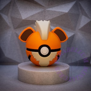 Op de afbeelding: Een oranje en witte Arcanine-thema Poké Ball replica, met bruine oren en een witte manen. De bal staat op een grijze cilindrische sokkel tegen een getextureerde grijze achtergrond.