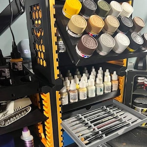 May include: Black and yellow model paint organiser with multiple shelves and a pull-out tray for brushes. The organiser holds various paint bottles and jars, with a small model tank visible on a lower shelf. The organiser is designed to keep painting supplies organised.
