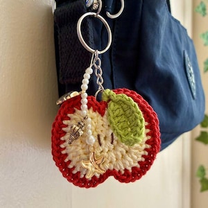Op de afbeelding: Een gehaakte appel sleutelhanger in rood, crème en groen, met schelp- en zeesterbedels en een parelsnoer. De sleutelhanger is bevestigd aan een marineblauwe tas. Een handgemaakt accessoire.