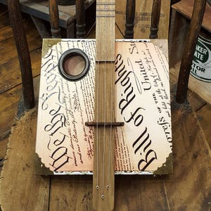 May include: A wooden cigar box guitar with a printed image of the Bill of Rights on the body. The guitar has a wooden neck and metal tuning pegs.