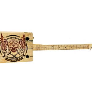 Puede incluir: Una guitarra de caja de puros de madera con un diseño de calavera y alas. La guitarra tiene un acabado de madera natural y el diseño es en rojo y marrón. El texto "HOMEMADE MUSIC" está impreso en la guitarra.