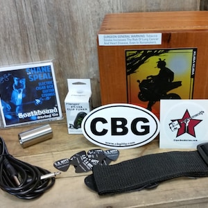 Może przedstawiać: Drewniana skrzynka na cygara z naklejką przedstawiającą osobę jadącą na motocyklu. Skrzynka zawiera zestaw strun do gitary, tuner do gitary, pasek do gitary, slide do gitary, plektron do gitary i kabel do gitary. Skrzynka ma również naklejkę z napisem "CBG" i naklejkę z napisem "Cigar Box Guitars".