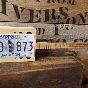 Puede incluir: Una guitarra de caja de puros de madera con una placa de matrícula de Mississippi unida al frente. La placa de matrícula dice "Mississippi" y "1GD 873" con la ciudad "Jackson" debajo del número. La guitarra tiene un mástil de madera y un cuerpo de madera clara.