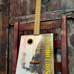 May include: A handmade cigar box guitar with a wooden neck and fretboard. The rectangular body features a vintage train illustration and the word "SOUTHBOUND" in yellow. The guitar rests on a wooden chair.