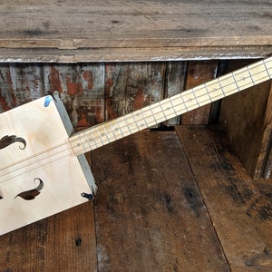 Puede incluir: Una guitarra de caja de puros de madera con un acabado de madera natural. La guitarra tiene un cuerpo rectangular con un agujero de sonido y un cuello largo con trastes. La guitarra está apoyada sobre una superficie de madera.