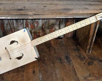 May include: A wooden cigar box guitar with a natural wood finish. The guitar has a rectangular body with a sound hole and a long neck with frets. The guitar is resting on a wooden surface.