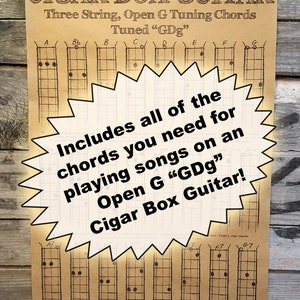 May include: A brown paper chart with a large yellow starburst in the center. The starburst has text that reads "Includes all of the chords you need for playing songs on an Open G "GDg" Cigar Box Guitar!" The chart shows diagrams for guitar chords, labeled with letters and numbers. The chart is titled "Cigar Box Guitar Three String, Open G Tuning Chords Tuned "GDg""