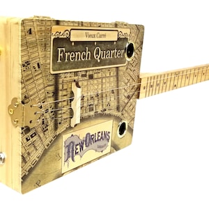 Puede incluir: Una guitarra de caja de puros de madera con un mapa del Barrio Francés de Nueva Orleans en la parte delantera. La guitarra tiene un mástil de madera y una clavija de afinación de metal. La guitarra es un instrumento hecho a mano.