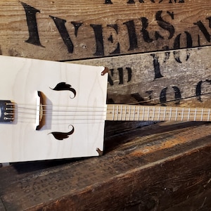 Puede incluir: Una guitarra de caja de madera blanca con un diapasón y clavijas de afinación. La guitarra está apoyada en una caja de madera con el texto "IVERSOND AND CO" impreso en ella.
