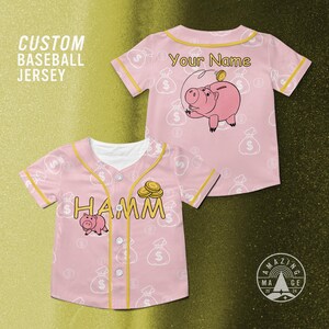 Puede incluir: Camiseta de b&eacute;isbol rosa personalizada con ribete amarillo y botones blancos. La camiseta presenta un gr&aacute;fico de cerdo de dibujos animados y la palabra "HAMM". El fondo tiene un patr&oacute;n de bolsas de dinero y s&iacute;mbolos de d&oacute;lar. El texto "Your Name" tambi&eacute;n es visible.