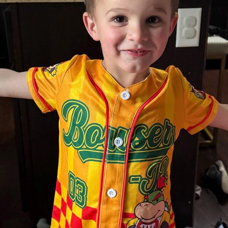 Może przedstawiać: Ż&oacute;łto-pomarańczowa koszulka baseballowa w paski z napisem Bowser Jr zieloną czcionką, numerem 03 w kolorze zielonym oraz grafiką postaci Bowser Jr. Koszula zapinana na guziki ma czerwone lam&oacute;wki, kr&oacute;tkie rękawy i białe guziki.