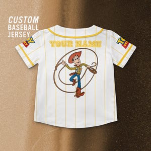 Może przedstawiać: Biała koszulka baseballowa z ż&oacute;łtymi prążkami i wykończeniami. Zawiera kresk&oacute;wkową grafikę kowboja i napis "YOUR NAME" na plecach. Koszulka ma r&oacute;wnież napis "CUSTOM BASEBALL JERSEY".