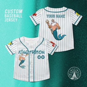 Puede incluir: Camiseta de béisbol blanca con finas rayas azules y una ilustración de dibujos animados del Rey Tritón. La camiseta tiene el texto "KING TRITON" y "00" en la parte delantera, y "YOUR NAME" en la parte trasera. Las mangas tienen ilustraciones de peces.