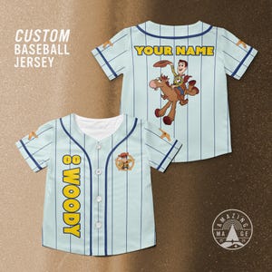 Puede incluir: Camiseta de b&eacute;isbol azul claro con rayas blancas verticales y ribetes azul marino. La camiseta presenta el texto "WOODY" en amarillo en la parte delantera y una imagen de dibujos animados de Woody montando a caballo. La parte posterior de la camiseta tiene el texto "YOUR NAME".