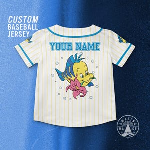 May include: White baseball jersey with yellow pinstripes, featuring a cartoon Flounder graphic and the text "YOUR NAME". The jersey has blue accents and the text "CUSTOM BASEBALL JERSEY".