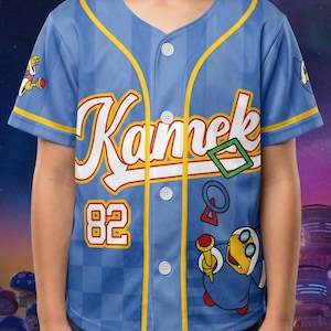 Puede incluir: Una camiseta de b&eacute;isbol azul claro con botones que presenta el nombre Kamek en letra blanca, el n&uacute;mero 82 en amarillo y rojo, y un gr&aacute;fico del personaje Kamek sosteniendo una varita. La prenda tiene ribetes amarillos y un patr&oacute;n de cuadros.