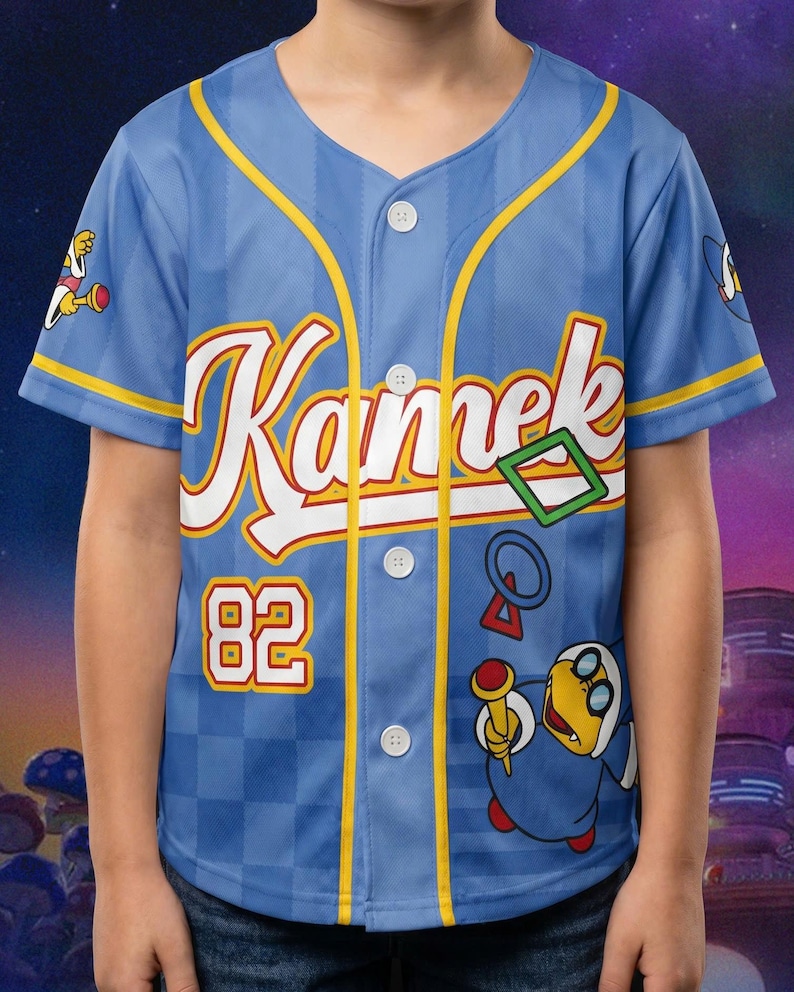 Puede incluir: Una camiseta de b&eacute;isbol azul claro con botones que presenta el nombre Kamek en letra blanca, el n&uacute;mero 82 en amarillo y rojo, y un gr&aacute;fico del personaje Kamek sosteniendo una varita. La prenda tiene ribetes amarillos y un patr&oacute;n de cuadros.
