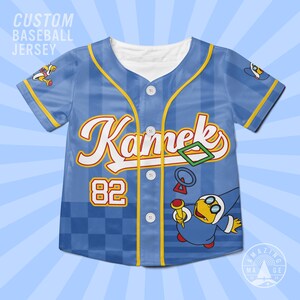 May include: A light blue baseball jersey featuring a checkered pattern at the bottom, yellow trim, and white buttons. The front displays the text Kamek in white with a yellow outline, the number 82, and a graphic of the character Kamek holding a wand.