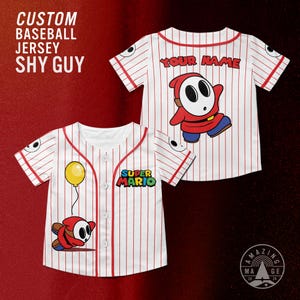May include: Two white baseball jerseys with red pinstripes and accents. One jersey features a Shy Guy character with a yellow balloon, and the other has the text "YOUR NAME" above a Shy Guy. The Super Mario logo is on one jersey. Text on the image reads "CUSTOM BASEBALL JERSEY SHY GUY".