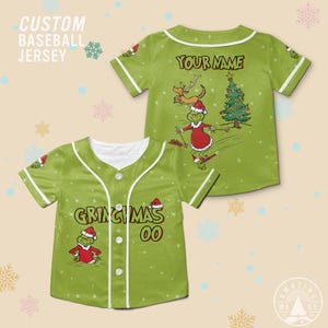 Könnte beinhalten: Grünes Baseball-Trikot mit weißer Paspelierung und Knöpfen. Auf der Vorderseite steht der Text "GRINCHMAS 00" und ein Cartoon-Grinch mit Weihnachtsmütze. Auf der Rückseite steht der Text "YOUR NAME" und ein Grinch beim Skifahren.
