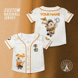May include: White baseball jersey with orange trim and vertical stripes. Features a cartoon character graphic and the text "YOUR NAME" on the back. The front displays the text "UP" and a cartoon graphic.