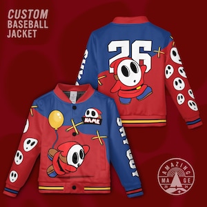 May include: Custom baseball jacket featuring a Shy Guy character design. The jacket is red and blue with a yellow balloon graphic, Shy Guy character, and the text "NAME". The sleeves have white Shy Guy face graphics. The back has the number 26 and the Shy Guy character.