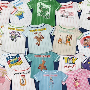 Puede incluir: Colecci&oacute;n de camisetas estilo b&eacute;isbol con varios personajes de Toy Story. Cada camiseta presenta un personaje diferente, rayas y el texto "YOUR NAME" encima del personaje. Los colores incluyen blanco, verde, azul y rosa.